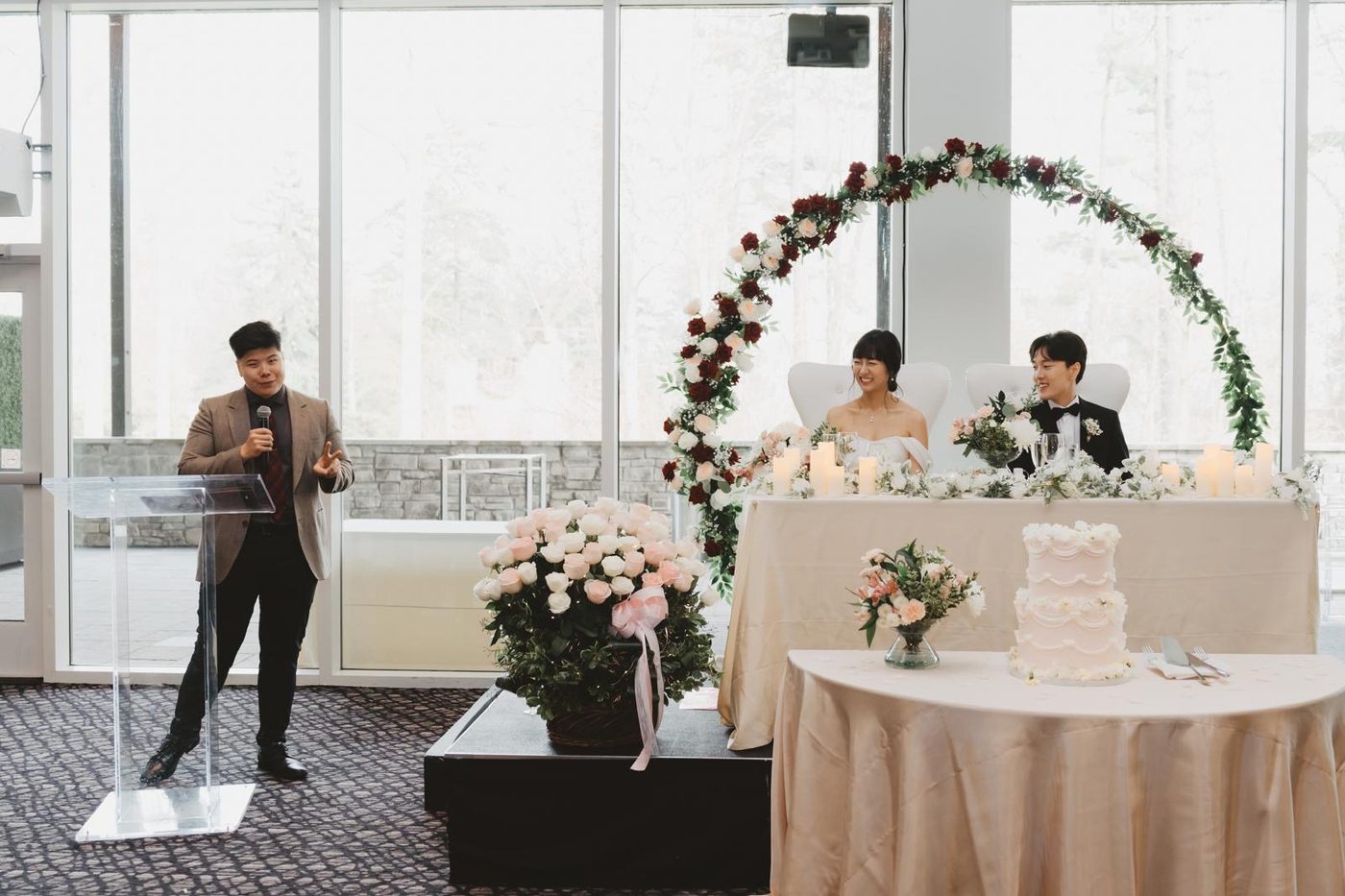 A full wedding reception room mid-toast, floral arch and cake table in view, Anthony hosting from the side.