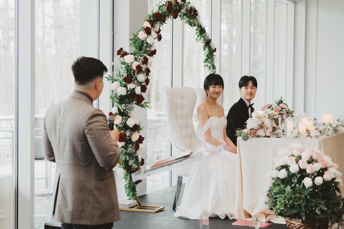 Anthony speaking to the newlyweds at their sweetheart table, framed by a rose and greenery arch.