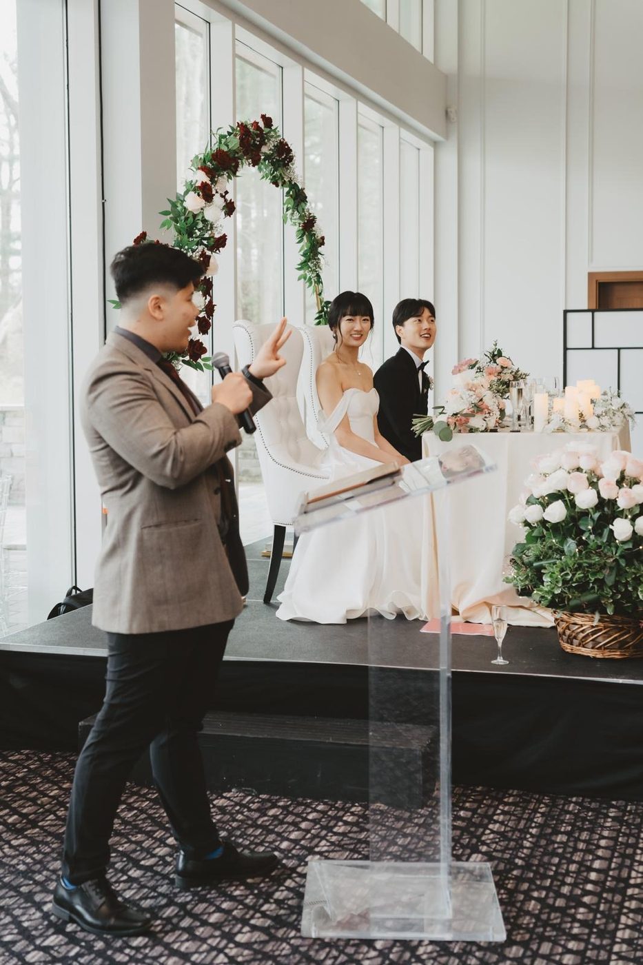 Anthony hosting a wedding reception, mic in hand, in front of the newlyweds' sweetheart table.